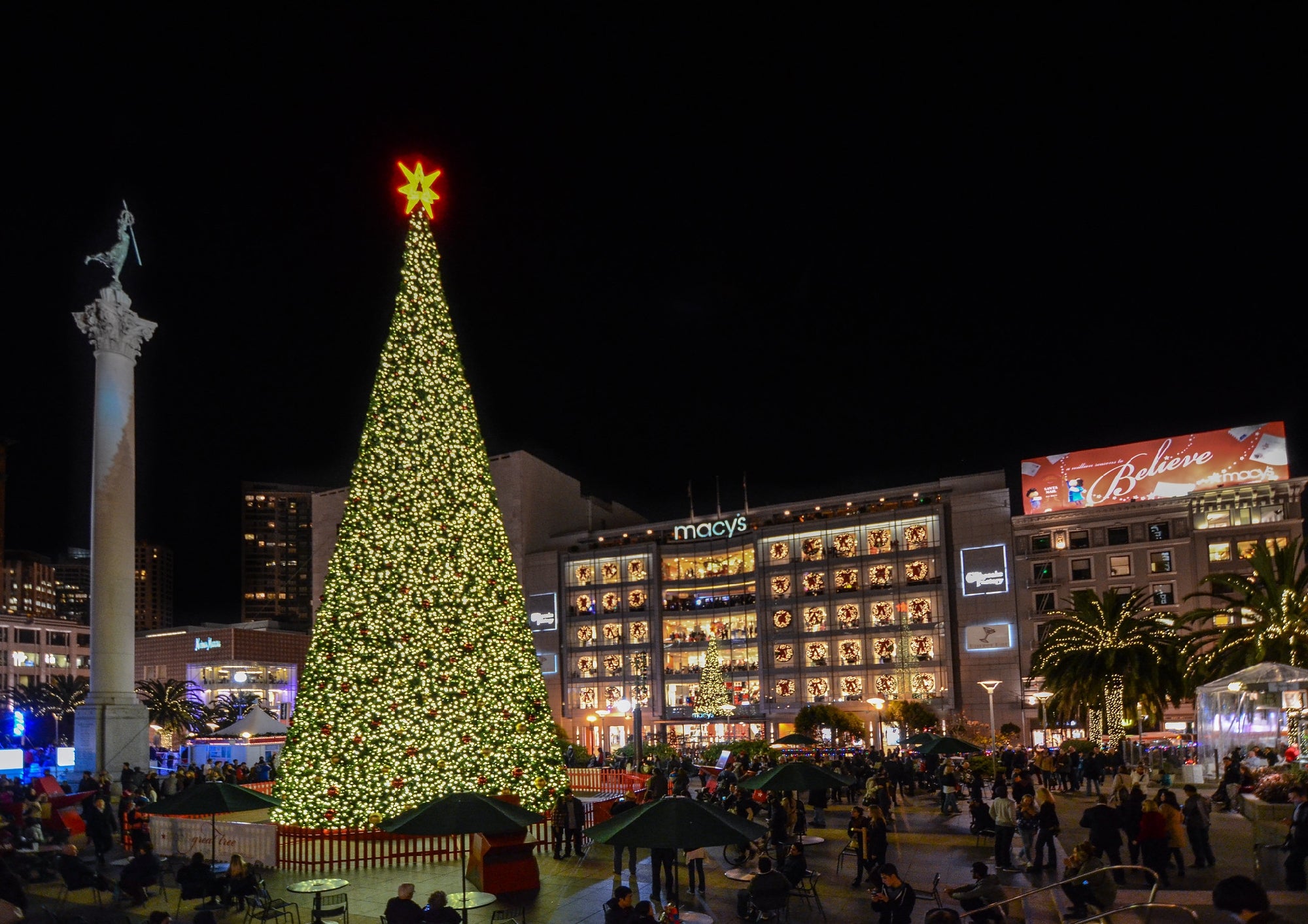 A large, decorated Christmas tree lit up at night in a busy city square, with crowds of people, surrounding buildings, and store signs visible in the background.