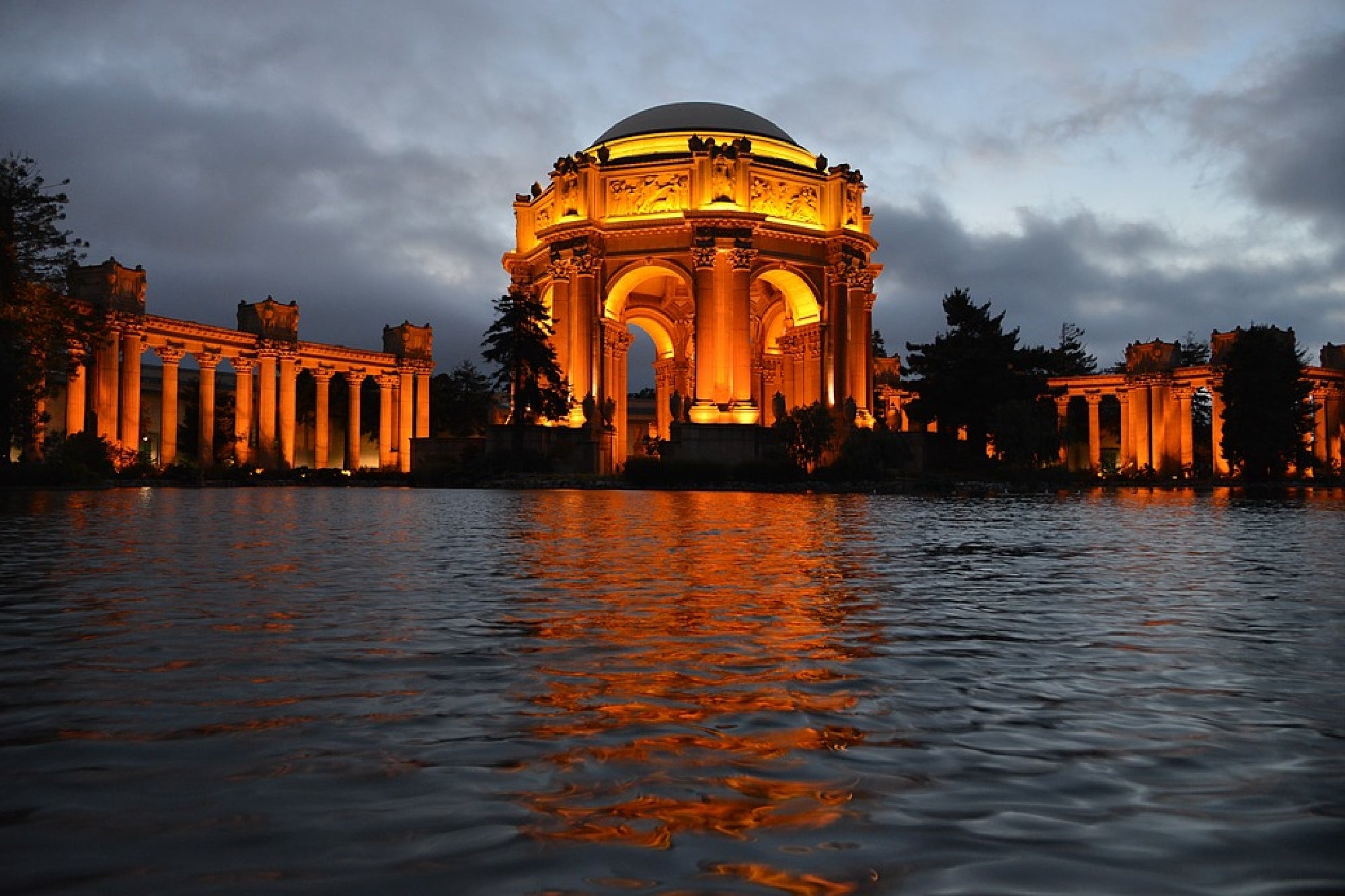 The illuminated Palace of Fine Arts in San Francisco is reflected in a pond at dusk, with dramatic clouds and trees in the background.