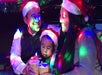 Three people wearing Santa hats sit closely together holding cups, illuminated by colorful lights in a festive setting.