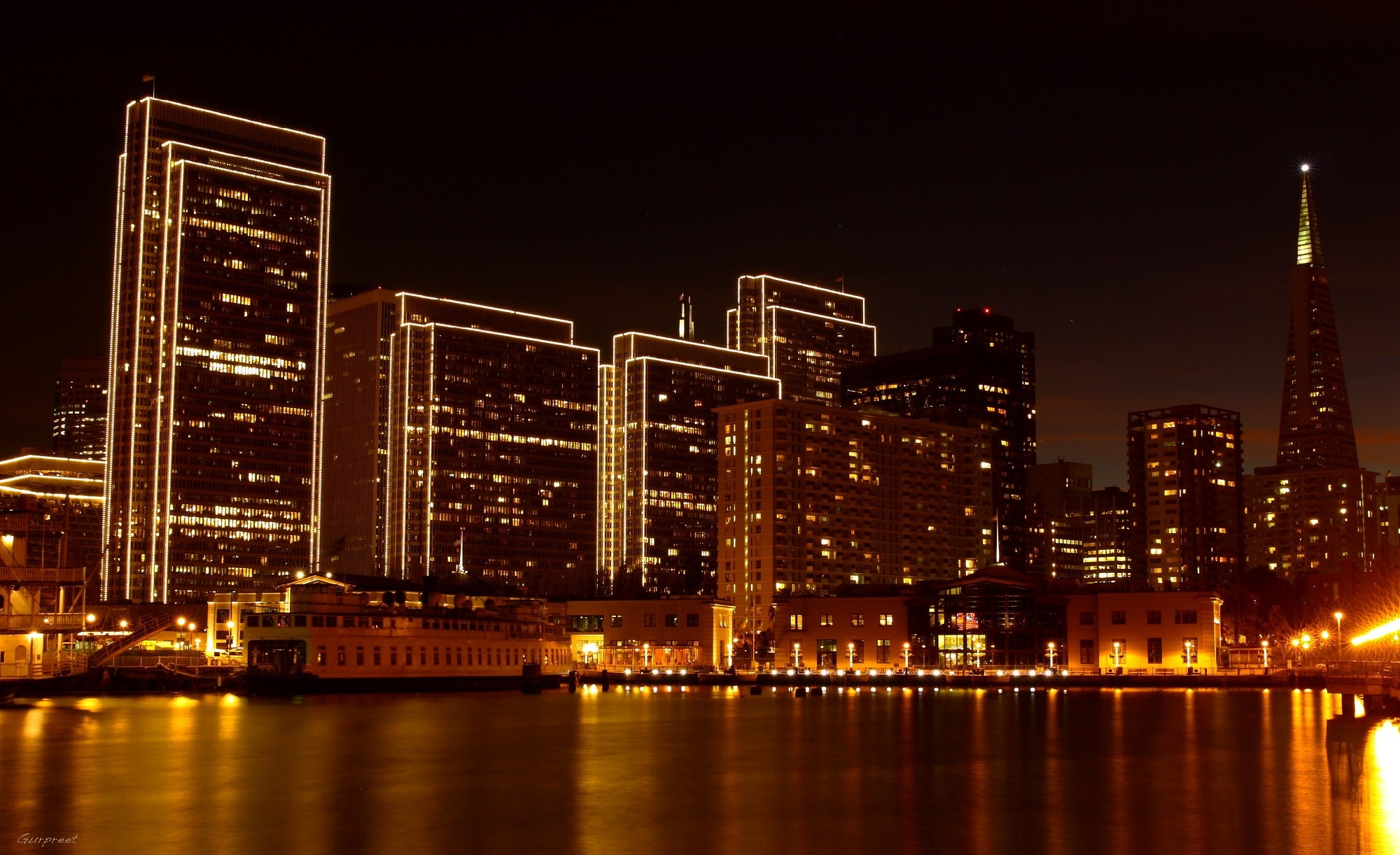 A nighttime cityscape shows illuminated high-rise buildings and the Transamerica Pyramid reflecting in calm waterfront waters.