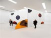 Two people stand near a white cube structure with circular colored windows and an open hinged orange panel, inside a gallery with white walls and ceiling lights.
