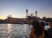 Two people on a boat look toward a baseball stadium at sunset, with stadium lights on and boats docked nearby.