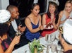 A group of people dressed festively sit around a table, smiling and clapping as one woman receives a plate with a slice of chocolate cake.