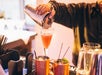 A bartender pours a pink cocktail from a shaker into a glass, with copper mugs and glasses on the bar counter.