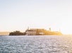 Alcatraz Island sits in the bay, surrounded by water, with prison buildings and a water tower visible under a bright, sunlit sky.