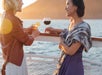Two women stand on a boat deck at sunset, holding drinks and talking, with water and a mountain in the background.