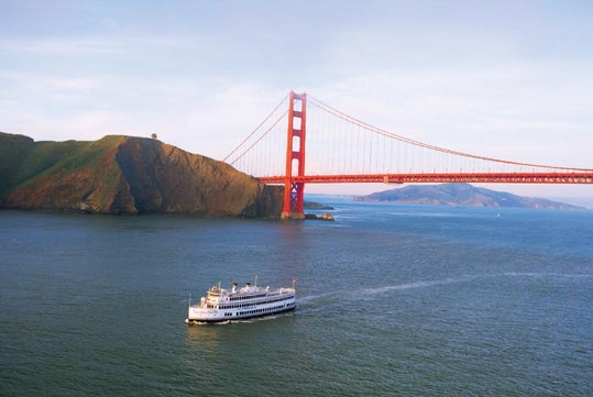 A white ferry boat sails in the bay near the Golden Gate Bridge, with green hills and blue water visible under a clear sky.