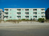 A four-story, light green beachfront building with multiple balconies, surrounded by sandy terrain and beachgrass, against a clear blue sky.