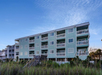 Three-story green beachfront apartment building with multiple balconies, surrounded by tall grass and a clear sky above.
