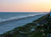 A serene beach scene at sunset with calm waves, sandy shore, and vegetation-covered dunes. The sky is a gradient of warm pastel colors.