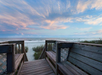 Wooden boardwalk leading to a sandy beach with gentle waves beneath a sky painted with pink and blue clouds.