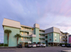 A pastel green, three-story residential building with balconies and palm trees in front. Several cars are parked in the lot. The sky is partly cloudy at sunset.