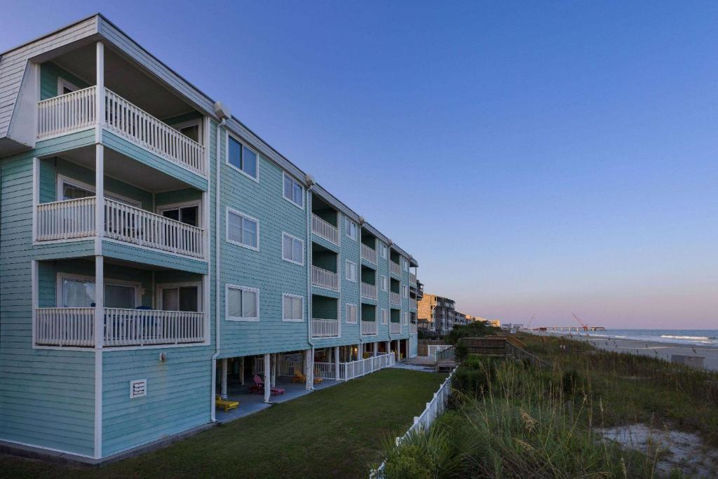 A four-story, light blue beachfront apartment building with balconies, adjacent to grass and dunes, overlooking the ocean at sunset.