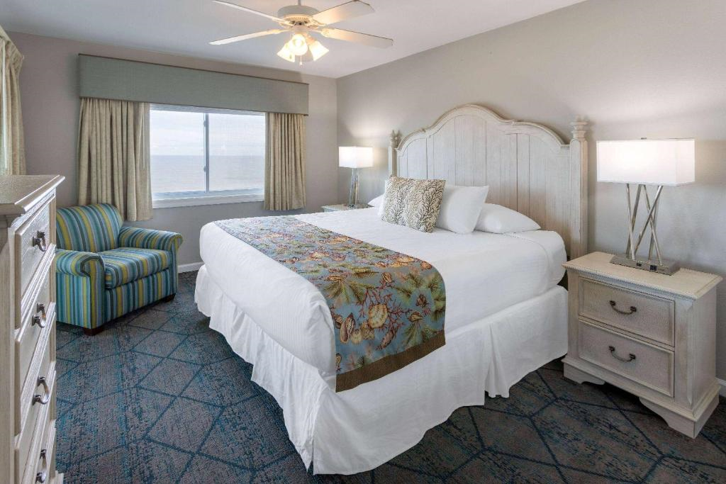 Room with a large bed adorned with floral runner, two bedside tables with lamps, a striped armchair, and a window overlooking the ocean. Ceiling fan above.