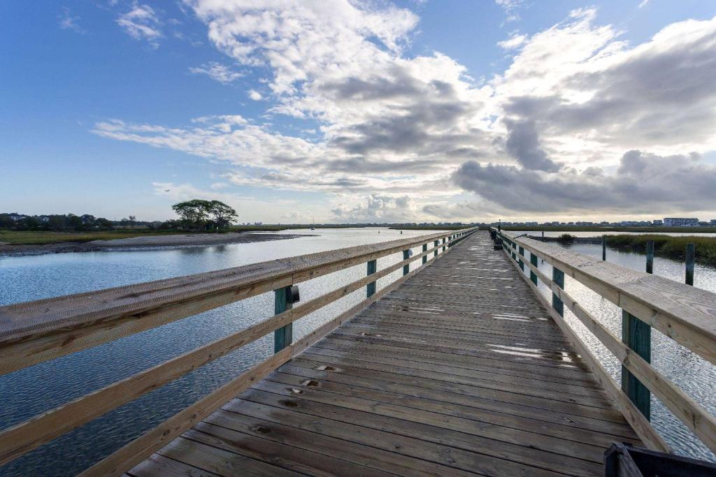 Long wooden bridge over calm water leading to a distant land, with a partly cloudy sky and scattered trees in the background.