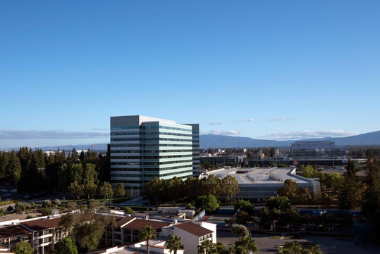 Aerial view of a modern office building surrounded by trees and smaller structures, with mountains visible in the background under a clear blue sky.