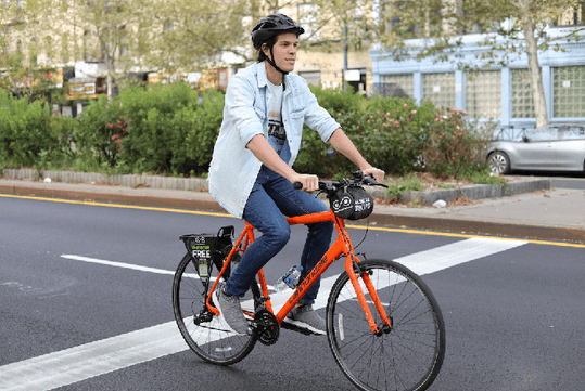 A man wearing a light blue jacket, jeans, and a black helmet while riding a red-orange bicycle down the street in Santa Monica.