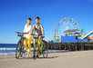 A man and woman wearing helmets posing with the bicycles in the sand near the ocean with Santa Monica Ferris Wheel in the background.