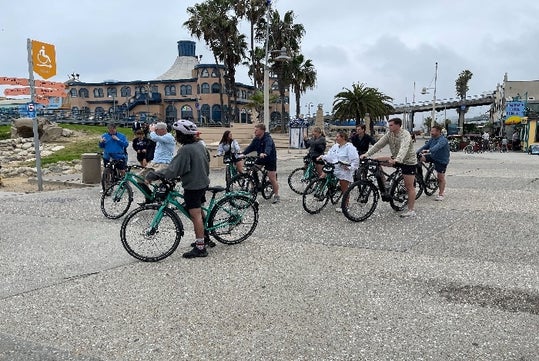 A group of tourists stopped on their bicycles and listening to their tour guide with a large building and palm trees in the background.