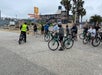 A group of cyclists gathers at a beachside path near a building and palm trees. One person wears a yellow vest and appears to be instructing the group.