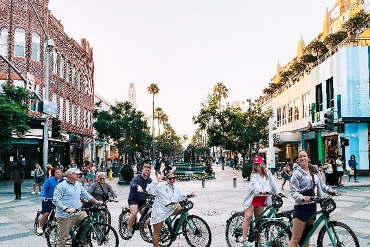 A group of people riding bicycles on a busy, tree-lined street with shops and buildings on either side.
