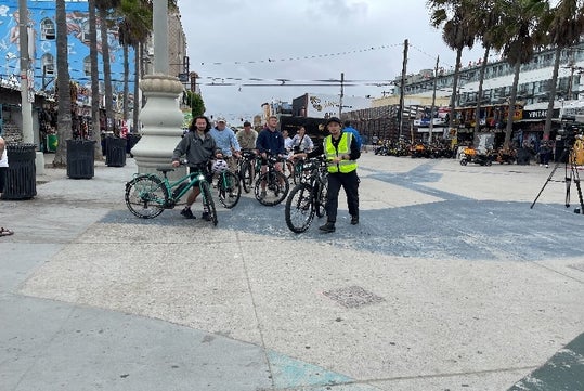 A group of people stand with bicycles on a wide promenade, surrounded by shops and palm trees on a cloudy day.