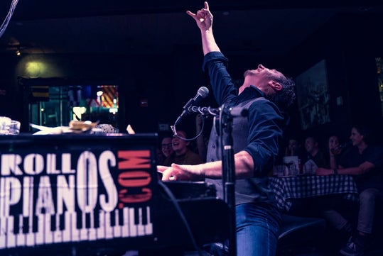 A musician performs energetically at a piano, with an enthusiastic crowd in the background.