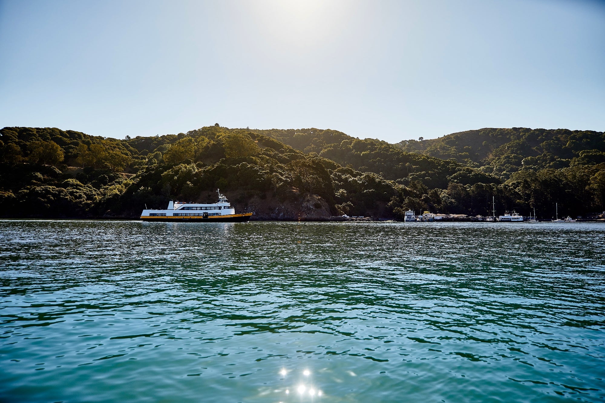 A yellow ferry sails on a calm body of water with tree-covered hills in the background under a clear sky.