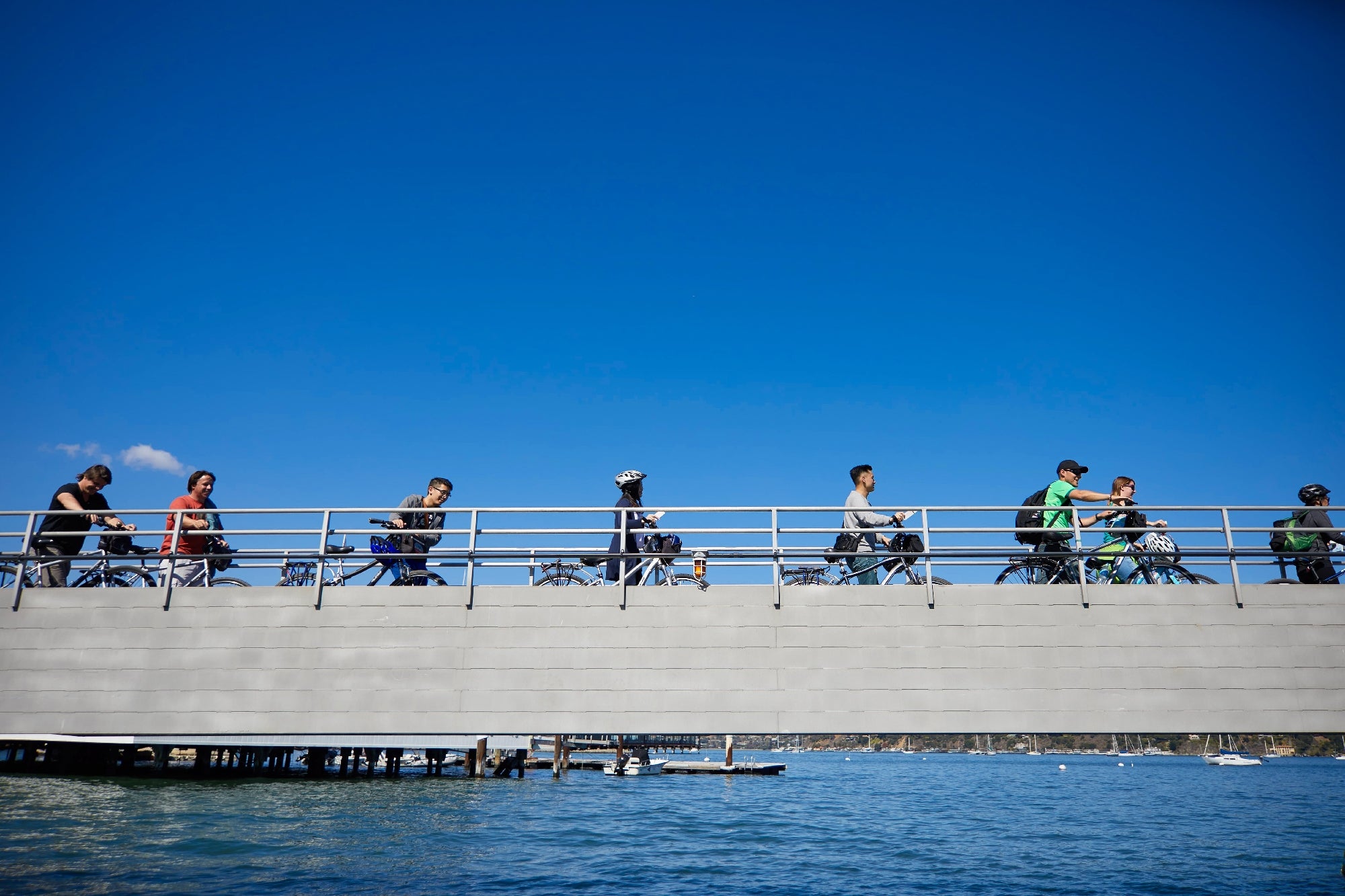 People ride bicycles across a bridge over water on a sunny day.