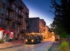 A streetcar with interior lights on travels along cobblestone tracks past old brick buildings at dusk in an urban area.