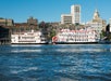 Two white riverboats with red trim docked along a waterfront, with historic brick buildings and modern structures in the background under a clear sky.