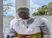 A woman wearing a green headwrap, a white ruffled blouse, and a yellow skirt stands outdoors near a white column, with trees and blue sky in the background.