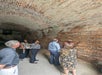A group of people listen to a guide speaking in a brick tunnel, with arched ceilings and dim lighting.