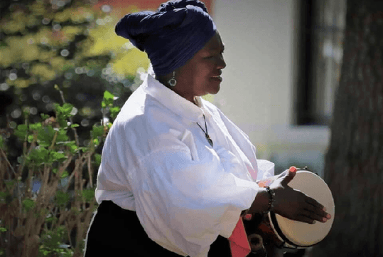 A person in a blue headwrap and white shirt plays a hand drum outdoors, with sunlight filtering through trees in the background.