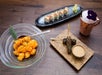 A glass bowl of sauced shrimp, a plate of sushi rolls with wasabi and ginger, skewered meat with sauce, and a copper cup with ice and a purple flower, on a wooden table.