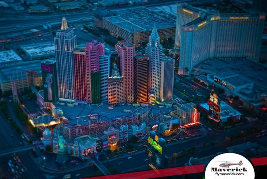 Aerial view of the New York-New York Hotel and Casino in Las Vegas at dusk, with colorful building lights and a visible roller coaster. Maverick Helicopters logo in lower right corner.