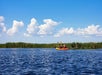 Two people paddle a yellow and red kayak on calm water, with green reeds and clouds in the background under a clear blue sky.