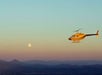 A yellow helicopter flies above a landscape with distant hills at sunset, with the moon visible in the sky.
