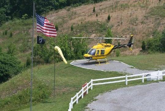 A yellow helicopter is parked on a helipad near a white fence, with an American flag and a windsock nearby, on a grassy hillside.