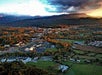 Aerial view of a small city surrounded by trees and fields, with mountains in the background under a partly cloudy sky at sunset.