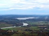 Aerial view of a rural landscape featuring a winding river, a large lake, scattered houses, fields, and distant hills under a cloudy sky.