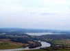 Aerial view of a winding river with surrounding fields, scattered houses, and distant hills under a mostly cloudy sky.