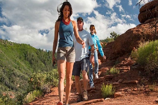 Three people hike along a rocky trail in a scenic outdoor setting with green hills, blue sky, and scattered clouds in the background.