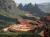 A view of red rock formations and layered mesas surrounded by green forest in a mountainous landscape under a partly cloudy sky.