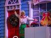 An older woman in festive clothing stands in front of a decorated house set, holding a whisk and smiling with arms outstretched.