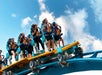 A group of people ride a roller coaster, secured in seats with harnesses, as the track tilts upward against a blue sky with clouds.