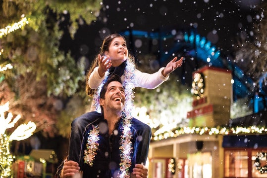 A man gives a girl a piggyback ride as they both look up and smile, surrounded by festive lights and decorations at night.