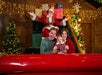 A family with a baby poses and smiles in front of Santa Claus, who sits with arms raised on a decorated stage with Christmas trees and lights.