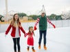 Three people ice skating outdoors, wearing winter clothing and holding hands. Roller coasters visible in the background.