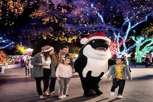 A family walks with a person in an orca costume wearing a Santa hat, surrounded by trees decorated with colorful holiday lights at night.